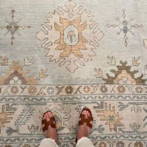 May include: A light blue and beige patterned rug with a central medallion design. The rug features intricate geometric patterns in shades of beige, brown, and blue. The rug is rectangular and appears to be made of woven material. The feet of a person wearing brown sandals are visible.