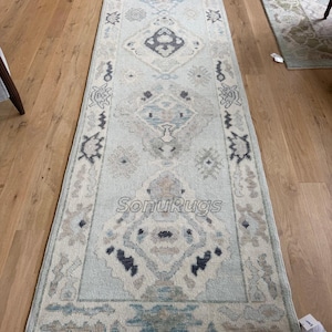 May include: A rectangular, light blue and beige patterned rug with a central diamond design and geometric borders. The rug is placed on a wooden floor, showcasing its intricate details and color palette. The text "SonuRugs" is visible on the rug.