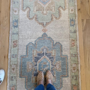 Puede incluir: Una alfombra rectangular de estilo vintage con un diseño de medallón central en tonos azules, verdes y beige. La alfombra presenta un patrón intrincado con elementos florales y geométricos. La alfombra está colocada sobre un suelo de madera.