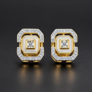 May include: A pair of gold-toned square earrings with a white center and a square clear stone. The earrings are surrounded by small, clear crystals. The background is black.