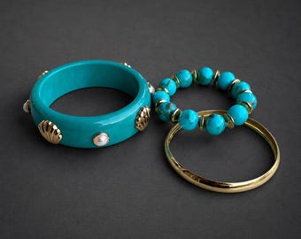 May include: Three bracelets on a dark background. A wide, teal bangle with gold seashell accents and small pearls. A beaded bracelet with turquoise beads and gold accents. A thin, gold bangle.