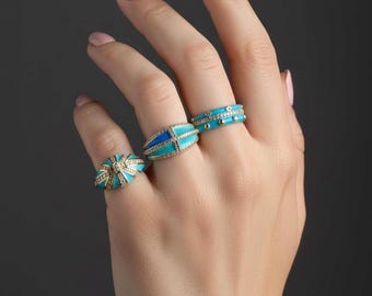 May include: A hand displays three rings with turquoise enamel and diamond accents. The rings feature various designs, including a band with small diamonds, an oval shape with a geometric pattern, and a dome shape with a starburst design.