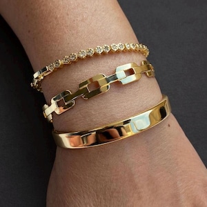 May include: A set of three gold-toned bracelets. The top bracelet features a delicate hexagonal link design with small embedded stones. The middle bracelet is a chunky chain link style. The bottom bracelet is a smooth, solid gold-toned cuff.