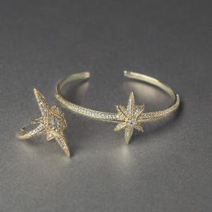 May include: A gold-toned jewelry set featuring a ring and a cuff bracelet. Both pieces are adorned with starburst designs and small, sparkling crystals. The bracelet has an open design, and the ring has a starburst design.