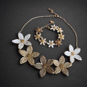 May include: A gold-tone chain necklace and matching bracelet set featuring five-petaled flowers crafted from textured white and tan fiber cord. Each flower center is accented with small gold beads, and the jewelry includes lobster claw clasps.