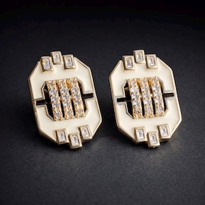 May include: A pair of gold-coloured earrings with a geometric design. Each earring has a white enamel base with rectangular and square clear crystals. The centre has three rows of small, clear crystals.