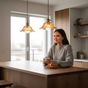 May include: Two pendant lights with amber-colored glass shades hang above a wooden kitchen island. The shades have a scalloped edge design. A person is seated at the island, holding a cup and looking out the window.