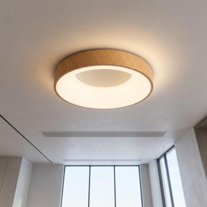 May include: A circular flush mount ceiling light fixture featuring a light-toned wood grain frame and a central white diffuser panel that emits a soft, warm glow, mounted directly against a smooth, flat white ceiling surface.