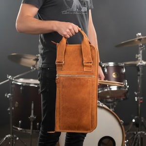 Könnte beinhalten: Eine braune Leder-Drumstick-Tasche mit Griff und Schultergurten. Die Tasche hat eine Reißverschluss-Vordertasche und eine strukturierte Oberfläche. Die Tasche wird vor einem Schlagzeug gehalten.