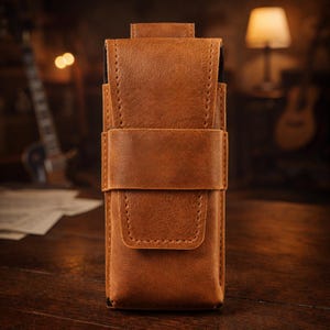 Puede incluir: Un estuche de cuero marrón con cierre de solapa y correa. El estuche es rectangular y tiene costuras visibles. El cuero tiene un aspecto texturizado. El estuche está diseñado para guardar un objeto pequeño, como un bolígrafo o una herramienta.