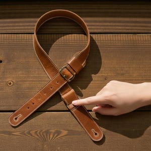 May include: A brown leather guitar strap with white stitching and a brass buckle. The strap is shown on a wooden surface. The strap has a classic design and is likely for an acoustic or electric guitar.