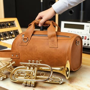 May include: A brown leather duffel bag with a zipper closure and top handles. A gold-colored trumpet and other brass instruments are in the foreground. The bag is being held by a person.