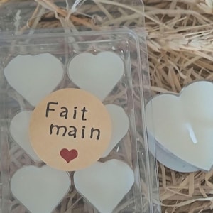 Puede incluir: Un recipiente de plástico transparente que contiene velas blancas en forma de corazón. Una pegatina redonda con las palabras "Fait main" y un corazón rojo está centrada en las velas. El fondo es un material natural y texturizado.