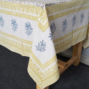 May include: A white tablecloth with a repeating pattern of blue floral designs and a yellow border. The tablecloth is draped over a wooden table, showcasing the intricate details of the fabric.