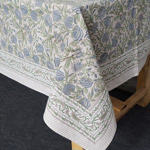 Puede incluir: Un mantel floral con un patrón repetitivo de flores azules y hojas verdes sobre un fondo crema. El borde presenta un diseño a rayas y elementos florales decorativos. El mantel está sobre una mesa de madera.