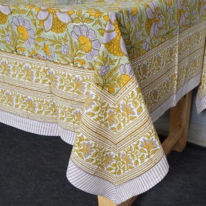 May include: A floral tablecloth with a repeating pattern of yellow and purple flowers on a light green background. The border features a detailed leaf design and a striped edge. The tablecloth is draped over a wooden table.