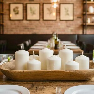 May include: A wooden bowl centerpiece holding white pillar candles of varying heights. The candles are arranged on a wooden table set for a meal, with plates, glasses, and silverware. The background shows a restaurant setting.