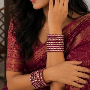 May include: A set of deep magenta bangles featuring intricate gold-toned metal detailing and embedded white stones. The bangles are arranged in stacks on both wrists, showcasing a consistent pattern of metallic accents and reflective stones.