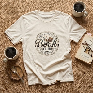 May include: A cream-colored short-sleeve t-shirt featuring a circular graphic with the text "What Happens at Book Club Stays at Book Club" surrounding an illustration of a small book and flowers, accompanied by two mugs of coffee and a book.