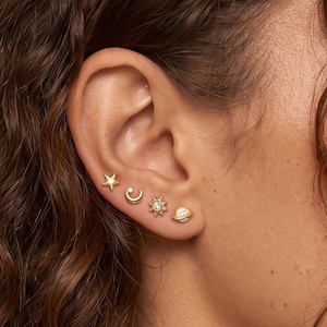 May include: A set of four small gold stud earrings worn in a single earlobe. The designs include a tiny star, a crescent moon, a sun with a central clear stone, and a small planet with a ring, all featuring a polished gold-tone metal finish.