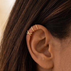 May include: A gold-toned ear cuff set consisting of three thin, delicate bands. The bottom band features a crossed design, while the two bands positioned above it are simple, smooth, circular hoops that sit along the upper cartilage of the ear.