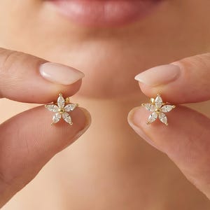 May include: A pair of gold stud earrings shaped like five-petaled flowers. Each earring features five marquise-cut clear stones arranged in a star-like pattern around a central point, creating a delicate floral design with a polished gold finish.
