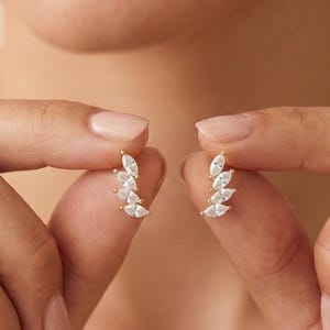 May include: A pair of gold-toned earrings, each featuring a curved row of four marquise-cut clear stones arranged in a leaf-like pattern. The earrings are held between fingers, showcasing the delicate metal setting and the faceted white stones.