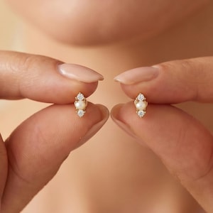 Puede incluir: Un pequeño pendiente de botón en oro con una perla blanca redonda central flanqueada por dos diminutas piedras claras brillantes dispuestas en línea vertical. El pendiente tiene un engaste de metal dorado pulido que sujeta la perla.