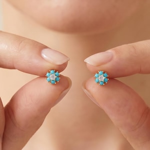 Pode incluir: Um par de brincos de tarracha com uma pedra preciosa central transparente rodeada por um grupo de seis pequenas pedras turquesas redondas e brilhantes, cravadas numa armação de metal dourado, criando um design floral em cada brinco.