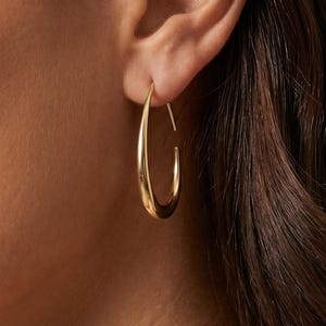 May include: A polished gold hoop earring with a teardrop shape. The earring features a smooth, reflective metallic surface and a tapered design that is wider at the bottom and narrows towards the top where it connects to the earlobe.