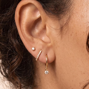 May include: A set of three gold-toned earrings consisting of a small stud with a clear stone, a thin bar-shaped earring encrusted with clear stones, and a small gold hoop earring featuring a single dangling clear stone charm.