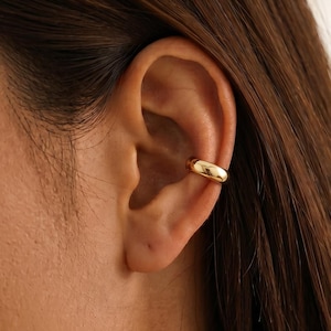 Può includere: Un ear cuff color oro, liscio e lucido, con un design a fascia semplice e arrotondato. Il gioiello è indossato sulla cartilagine superiore dell'orecchio e presenta una forma circolare continua e senza giunture che poggia sulla piega esterna.