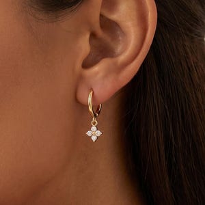Può includere: Un orecchino a cerchio in oro con un piccolo ciondolo a forma di fiore a quattro petali. Il ciondolo è incastonato con quattro minuscole pietre chiare a taglio rotondo disposte a motivo floreale, sospeso a un sottile cerchio in oro lucido.