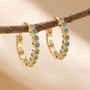 May include: A pair of gold hoop earrings featuring a row of small, round, bright blue turquoise-colored stones set along the front edge. Each hoop is stamped with the text S925 on the inner side, indicating the metal composition of the jewelry.