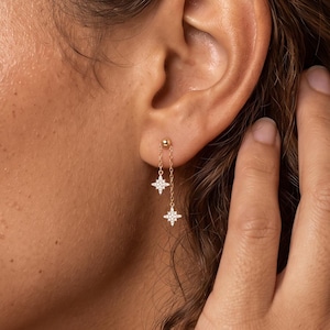 May include: A gold-toned earring featuring a small stud connected by a delicate chain to two dangling star-shaped charms. Each star charm is encrusted with tiny, clear, sparkling stones, creating a refined and symmetrical design that hangs vertically from the earlobe.