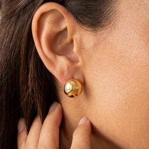 May include: A polished gold-tone spherical stud earring with a smooth, reflective surface. The earring is circular in shape and features a simple, classic design that sits flush against the earlobe, showcasing a bright, metallic yellow finish.