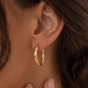 Könnte beinhalten: Ein polierter Gold-Creolen-Ohrring mit einer glatten, abgerundeten Oberfläche und einem kreisförmigen Design. Der Ohrring verfügt über ein reflektierendes Metallic-Finish, das das Licht einfängt und für einen hellen, klaren Look sorgt. Ein klassisches Schmuckstück.