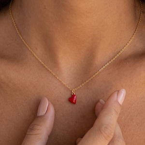 Op de afbeelding: Een delicate goudkleurige ketting met een kleine, glanzende rode hartvormige hanger. De hanger hangt centraal aan de dunne metalen ketting die op het sleutelbeen rust, wat zorgt voor een eenvoudig en verfijnd sieraad als accessoire.