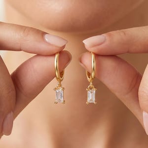 May include: A pair of gold hoop earrings, each featuring a small, clear, rectangular baguette-cut gemstone dangling from the bottom. The hoops are thin and circular, with the clear stones catching the light, creating a simple and elegant appearance.