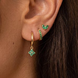 May include: A set of three gold-toned earrings featuring green stones. The set includes a small hoop earring with a dangling four-petal flower charm, a small huggie hoop earring with embedded green stones, and a small stud earring with two green stones.