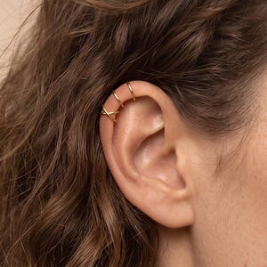 May include: A set of three thin gold-toned ear cuffs worn on the upper cartilage. The design consists of two plain circular bands and one band featuring a delicate crisscross detail, all positioned along the outer edge of the ear helix.