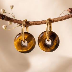 Puede incluir: Un par de pendientes de aro dorados con colgantes circulares de piedra ojo de tigre pulida. Cada pendiente consta de un aro de metal dorado liso que suspende una piedra en forma de donut con bandas características de color marrón y dorado.