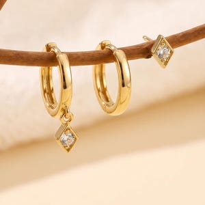 May include: A set of three gold-toned earrings consisting of two hoop earrings and one stud earring. One hoop features a small, diamond-shaped charm with a clear stone, while the stud earring also displays a matching diamond-shaped clear stone.