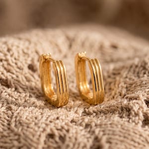Puede incluir: Un par de pendientes de aro dorados con forma rectangular y una textura acanalada de líneas múltiples en la superficie exterior. Los pendientes cuentan con un mecanismo de cierre de bisagra y un acabado metálico pulido y brillante.