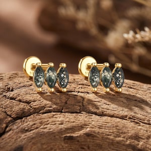 Könnte beinhalten: Ein Paar Ohrstecker aus Gold, jeweils mit drei im Marquise-Schliff facettierten, dunkelgrünen Moosachat-Steinen in horizontaler Anordnung. Die Ohrringe verfügen über einen flachen, runden Verschluss und eine Fassung aus poliertem Gold.