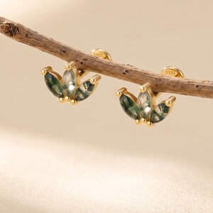 May include: A pair of gold stud earrings featuring three marquise-cut moss agate stones arranged in a fan shape. Each earring is crafted with a gold setting and includes a small S925 hallmark stamp on the back, displayed resting on a thin twig.