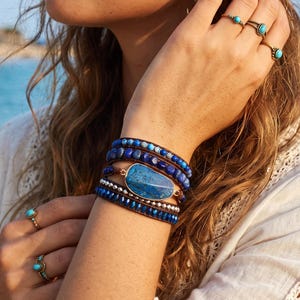 May include: A multi-strand wrap bracelet featuring blue lapis lazuli beads, small metallic silver spacers, and a large, central, irregular blue stone set in a copper-toned frame. Four rings with small blue stones are worn on the fingers.