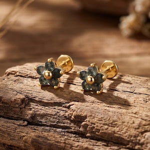 May include: A pair of gold stud earrings shaped like five-petaled flowers. Each earring features dark blue-grey stones set in the petals with a central gold bead. The posts are visible on the back, and the metal is stamped with 925.
