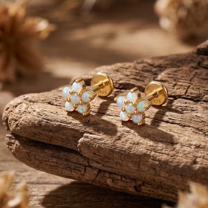May include: A pair of gold-toned stud earrings featuring a flower-shaped design. Each earring is composed of six small, round, iridescent white opals arranged in a circular pattern. The metal posts are clearly marked with the engraving S925.