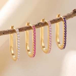 May include: Four gold-toned oval hoop earrings hanging from a thin wooden branch. Each earring features a row of small, sparkling gemstones along the front edge, with colors ranging from clear white to bright pink, light pink, and deep purple.
